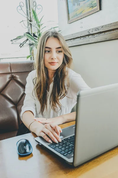 Young lady browsing the Internet at the cafe. Girl in a cafe with a laptop.Brunette girl with a gadget in the office. A woman is looking into a computer. Girl in a white shirt. Girl 18 or 19 years old