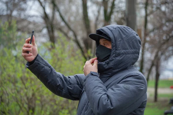 A man wearing a black respiratory mask on his face. Coronavirus. A guy in the spring on the street in a respiratory mask to protect against infection virus. A man photographs himself on a smartphone.