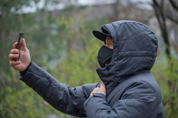 A man wearing a black respiratory mask on his face. Coronavirus. A guy in the spring on the street in a respiratory mask to protect against infection virus. A man photographs himself on a smartphone.