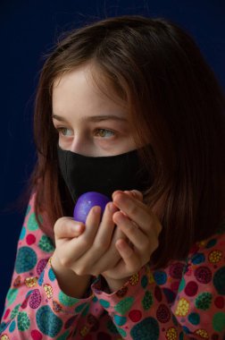 Mutlu Paskalyalar. Kız ve mor renkli yumurtalar. Paskalya gelenekleri. Ergen. Şarlatan bir kız. Puantiyeli elbise giymiş bir çocuk. Tatiller, bahar. Coronavirus, mavi arka planda maskeli bir kız..