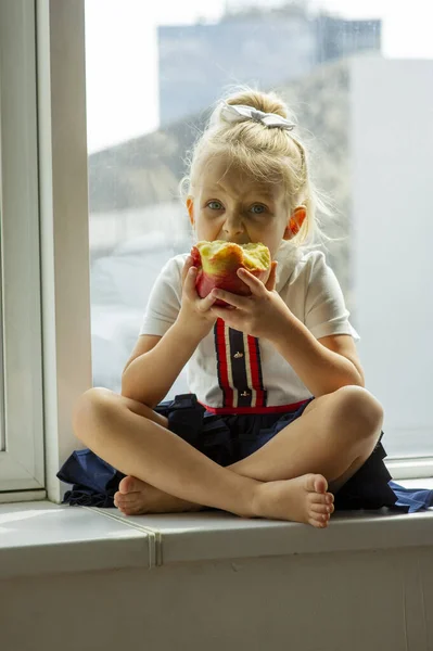 ブロンドの女の子は家に座って窓を見て赤いリンゴを持ってた 健康食品の概念 小さな女の子が窓でリンゴを食べている 女の子4または5歳でドレスにリンゴ — ストック写真