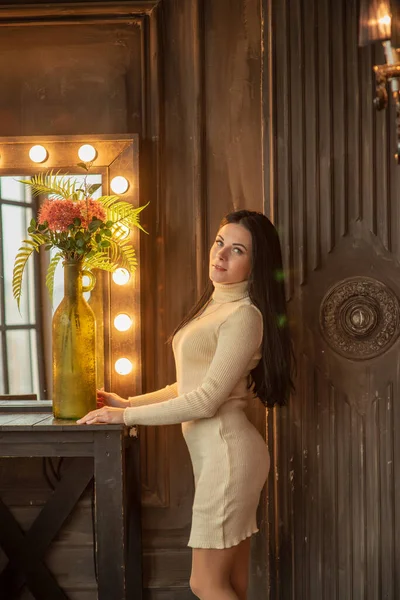 美しいエレガントなブルネットが背の高い化粧鏡の前に立っています 女の子が化粧台に立っている 白いニットのドレスを着た若い女の子の肖像 — ストック写真