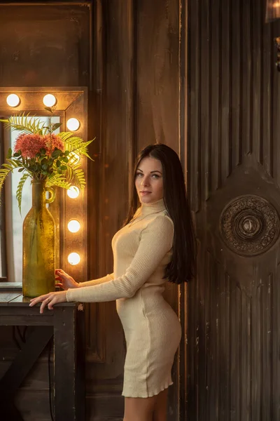 美しいエレガントなブルネットが背の高い化粧鏡の前に立っています 女の子が化粧台に立っている 白いニットのドレスを着た若い女の子の肖像 — ストック写真