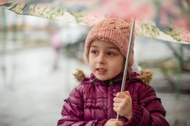 Dışarıda şemsiyenin altında küçük bir kız, yağmurlu bir gün. Küçük kız yağmurun altında şemsiyeyle yürür. Yağmurlu bir günde pembe şapkalı ve mor ceketli bir çocuk.