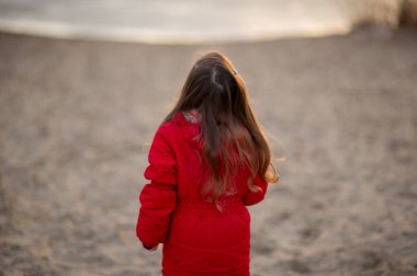 Kumsalda kumsalda yürüme yolu akşam güneşinde. Nehrin sonunda, kırmızı ceketli küçük bir kız. Eski model. Kırmızı ceketli küçük bir kız doğada yürüyor. Kum nehri. Portre