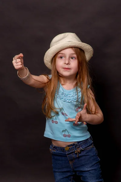 Beautiful girl doing different expressions: arms raised. Little girl on a black background in jeans. Portrait of a child. The girl in the hat. Emotional child.
