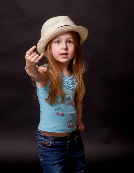 Beautiful girl doing different expressions: arms raised. Little girl on a black background in jeans. Portrait of a child. The girl in the hat. Emotional child.