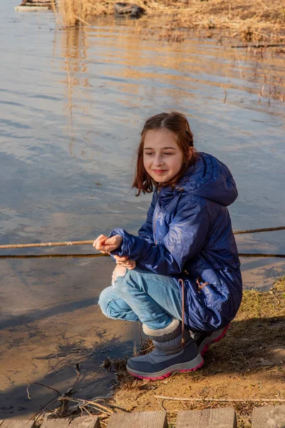 A girl in a light sheepskin coat stands on the bank of the river on a sunny day. Girl 9 years old in a sheepskin coat by the river. Girl 9 years old or 10 years old. Nature, child, schoolgirl.Teenager