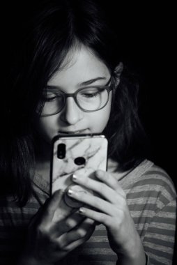 9 yaşında, siyah arkaplanlı, portre şeklinde bir çocuk portresi. Siyah arka planda 9 yaşında bir kız. Telefonlu bir kız. Akıllı telefonu olan bir çocuk. Gözlüklü genç.