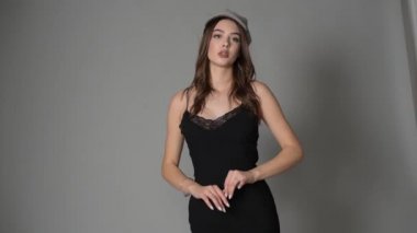 Göz kamaştırıcı genç kadın gri duvarda dalgalı saçlarıyla şık bir elbise giyiyor. Siyah elbiseli ve gri şapkalı bir kız elini öne uzattı. Kahverengi saçlı güzel bir genç kız. portre