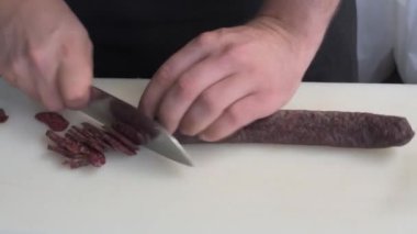 Açık havada bir adam kuru sosis dilimliyor. Bir adamın İspanyol usulü domuz sucuğu dilimlerini kestiği yakın plan. Kesme tahtasına bıçak saplanmış bir domuz sosisi.