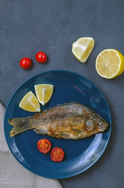 Smažené chutné rybí kapr s koření na modrém talíři na šedém pozadí zdobené plátky rajčat a citronu s nožem a vidličkou. kopírovat prostor. — Stock fotografie