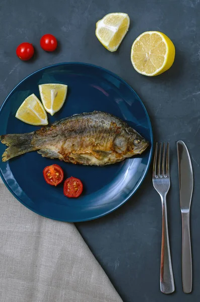 Smažené chutné rybí kapr s koření na modrém talíři na šedém pozadí zdobené plátky rajčat a citronu s nožem a vidličkou. svislý rámec. — Stock fotografie