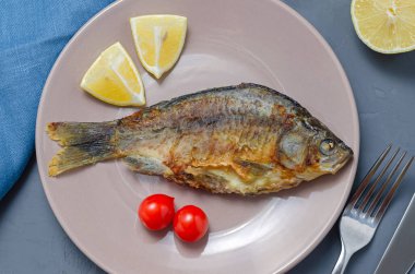 Kızarmış iştah açıcı balık sazan balığı, bej bir tabakta baharatlı, gri bir zemin üzerinde domates ve limon dilimleri ile süslenmiş bıçak ve çatalla. sağlıklı yeme kavramı.