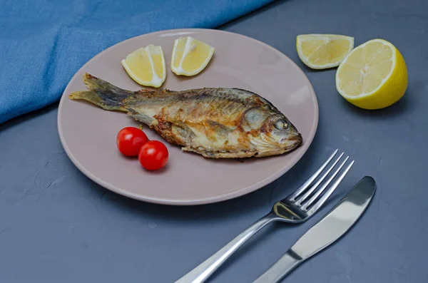 Crapul crucian de pește apetisant prăjit cu condimente pe o placă bej pe un fundal gri decorat cu felii de roșii și lămâie cu un cuțit și furculiță. Conceptul alimentar sănătos . — Fotografie, imagine de stoc