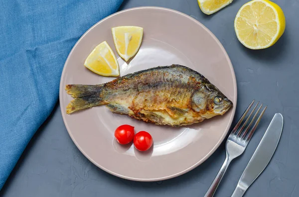 Crapul crucian de pește apetisant prăjit cu condimente pe o placă bej pe un fundal gri decorat cu felii de roșii și lămâie cu un cuțit și furculiță. Conceptul alimentar sănătos . — Fotografie, imagine de stoc