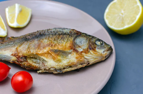 Crapul crucian de pește apetisant prăjit cu condimente pe o placă bej pe un fundal gri decorat cu felii de roșii și lămâie. Dieta mediteraneană. Conceptul alimentar sănătos . — Fotografie, imagine de stoc
