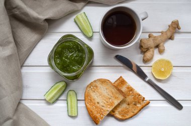 Yeşil salatalık reçeli şeffaf bir kavanozda, salatalık dilimleri ve kızarmış ekmekle zencefil, bir bıçak ve hafif ahşap arka planda tekstil peçetesi. Sağlıklı beslenme konsepti. flexitarian.