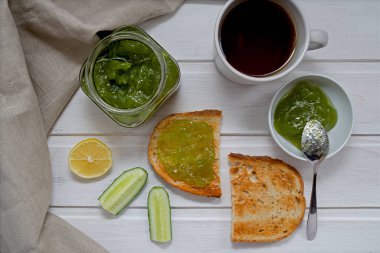 Yeşil salatalık reçeli şeffaf bir kavanozda, salatalık dilimleri ve kızarmış ekmekle zencefil, bir bıçak ve hafif ahşap arka planda tekstil peçetesi. Sağlıklı yeme kavramı.