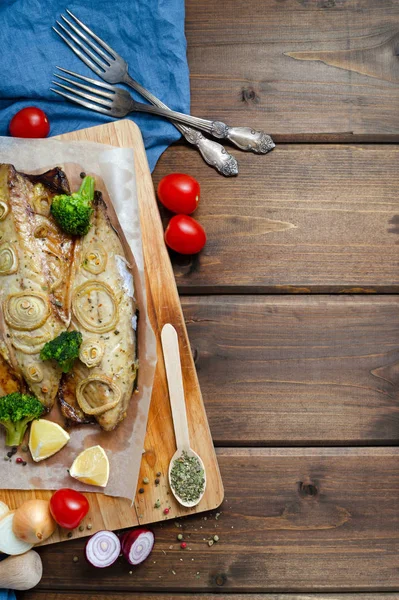 Closeup tender baked mackerel in the oven with rings of chopped onion, broccoli, tomatoes and lemon on dark wooden background with blue napkin and forks. keto and flexitarian diet and healthy eating. — Stock fotografie