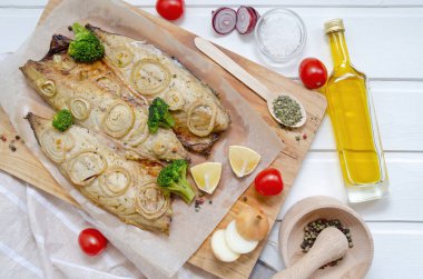 tasty mackerel fish with lemon, sliced on filet with tomatoes, onion rings, pepper, herbs, olive oil and broccoli in baking paper on a cutting board on a white wooden background