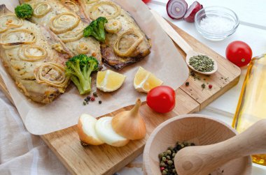 fresh baked tasty mackerel fish with lemon, sliced on filet with tomatoes, onion rings, pepper, herbs, olive oil and broccoli in baking paper on a cutting board on a white wooden background