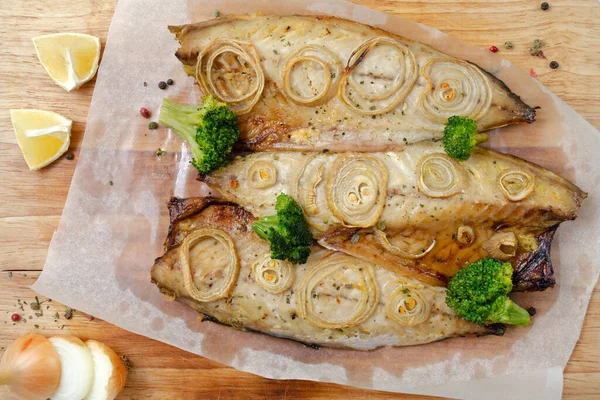 Closeup fresh tender baked mackerel with rings of chopped onion, broccoli and lemon on a dark wooden cutting board in baking paper. keto and flexitarian diet and healthy eating. — Stock Photo, Image