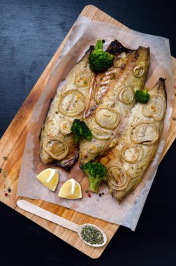 fresh tender baked mackerel with chopped onion, broccoli and lemon rings on a black slate background on a wooden cutting board in baking paper. keto and flexitritis diet and healthy eating. clipart