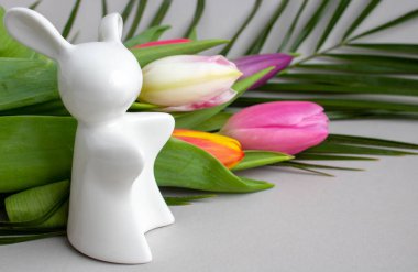 Beyaz seramik tavşan ve beyaz arka planda yeşil yapraklı çok renkli laleler.