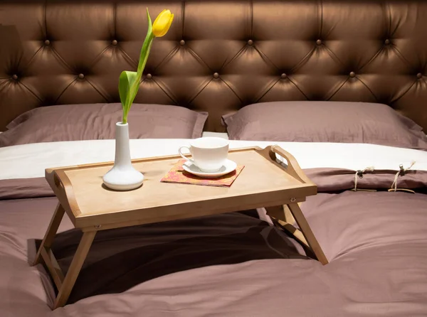 Wooden tray of coffee and candles with flowers on bed. Brown bedding sheets with striped blanket and pillow. Breakfast in bed. Hygge concept.