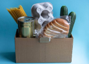 Ürünlerin eve teslimi. Pandemic sırasında insanlara yardım etmek. Mavi bir arka planda bir kutu yiyecek duruyor. Spagetti, salatalık, kavanozda tushonka, ekmek ve yumurtalı bir kap..