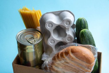 Ürünlerin eve teslimi. Pandemic sırasında insanlara yardım etmek. Mavi bir arka planda bir kutu yiyecek duruyor. Spagetti, salatalık, kavanozda tushonka, ekmek ve yumurtalı bir kap..