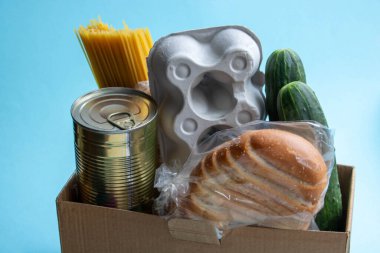 Ürünlerin eve teslimi. Pandemic sırasında insanlara yardım etmek. Mavi bir arka planda bir kutu yiyecek duruyor. Spagetti, salatalık, kavanozda tushonka, ekmek ve yumurtalı bir kap..