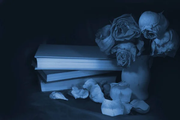 wilted rose flowers on a dark blue background, in a wooden vase, next to a stack of books and fallen rose petals, aged photo