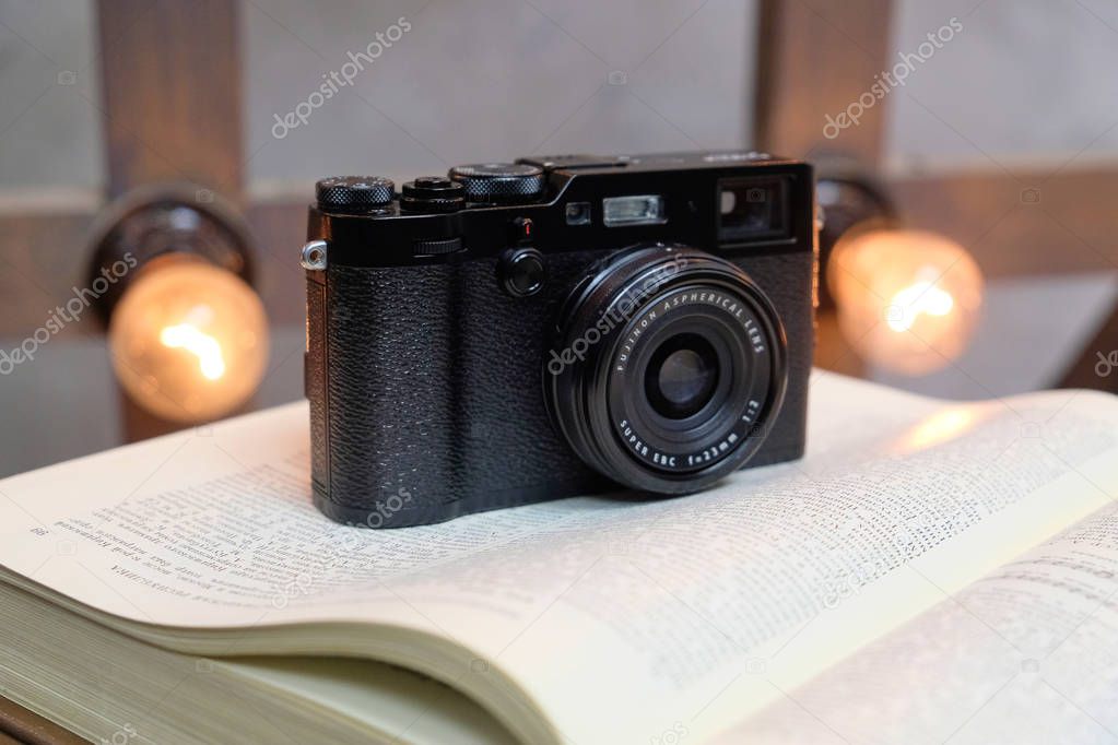 Belarus, the city of Minsk, st. Kuybysheva, Matryoshka Studio - November 30, 2017: a black Fujifilm X-100F camera is standing on an open book in Russian against the background of burning light bulbs t