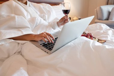 Şarap kadehi tutan ve lüks bir odada dizüstü bilgisayar oynayan genç bir kadının eli..