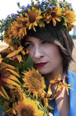 Buğday tarlasında duran genç bir kız mavi elbisenin içinde sarı bir sopa ile. Kafasında kır çiçeklerinden bir çelenk ve ayçiçekleri var..