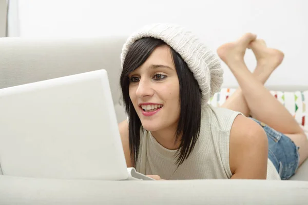 Junge brünette Studentin liegt auf der Couch mit einem Laptop — Stockfoto