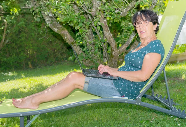 Mujer casual sentada en la silla de cubierta en el jardín con el ordenador portátil — Foto de Stock