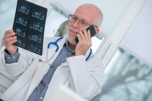 Volwassen arts kijken x-ray foto en via telefoon — Stockfoto