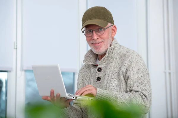 自宅のノート パソコンを使用してキャップを持つ年配の男性 — ストック写真