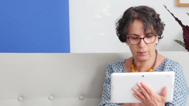 成熟的黑发女人 坐在沙发上使用一款平板电脑 — 图库视频影像