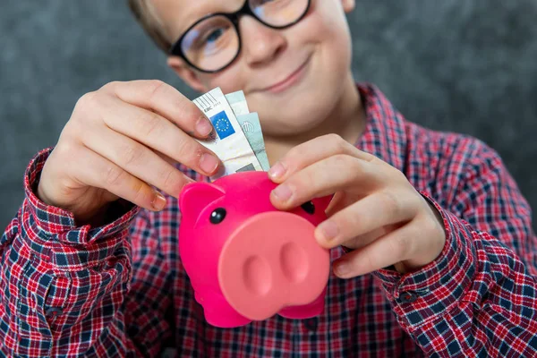 微笑带着眼镜的小男孩把钱投入一个存钱罐 — 图库照片