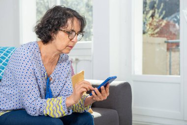 Kanepede otururken akıllı telefon kullanan neşeli, esmer yaşlı kadın.