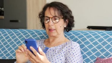 Kanepede otururken akıllı telefon kullanan neşeli, esmer yaşlı kadın.