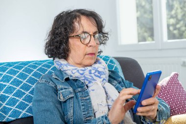 Kanepede otururken akıllı telefon kullanan neşeli bir esmer kadın.