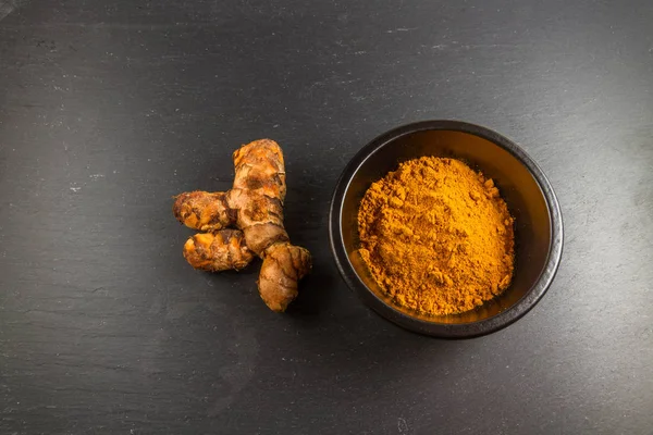 Radice di curcuma con ciotola di polvere su sfondo ardesia scura — Foto Stock