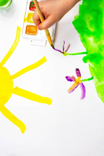 Dziecięcy ręka z pędzel maluję purpurowy kwiat na biały prześcieradło z zielony trawa i z żółty słońce. Farby akwarelowe i pędzle do malowania. Kreatywność dzieci. — Zdjęcie stockowe
