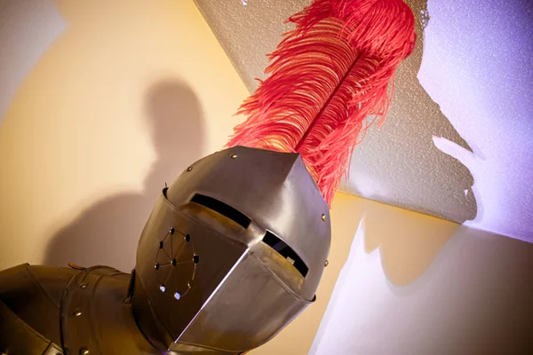 A suit of armor in a spotlight in a white room with red feather cap