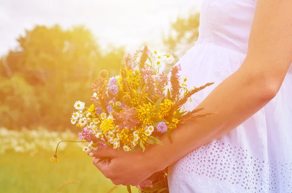Jovem grávida fica no campo abraçando o estômago à mão com um buquê de flores silvestres . — Fotografia de Stock